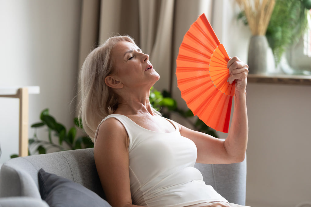 Stressed annoyed old senior woman using waving fan suffer from overheating, summer heat health hormone problem, no air conditioner at home sit on sofa feel exhaustion dehydration heatstroke concep